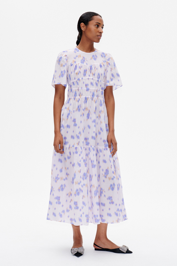 Anissa Dress Purple Fleur Fanee A mid-calf length dress with an A-line silhouette, elasticated panels through the body for a flattering look, and button closure with keyhole opening at the back of the neck - model image